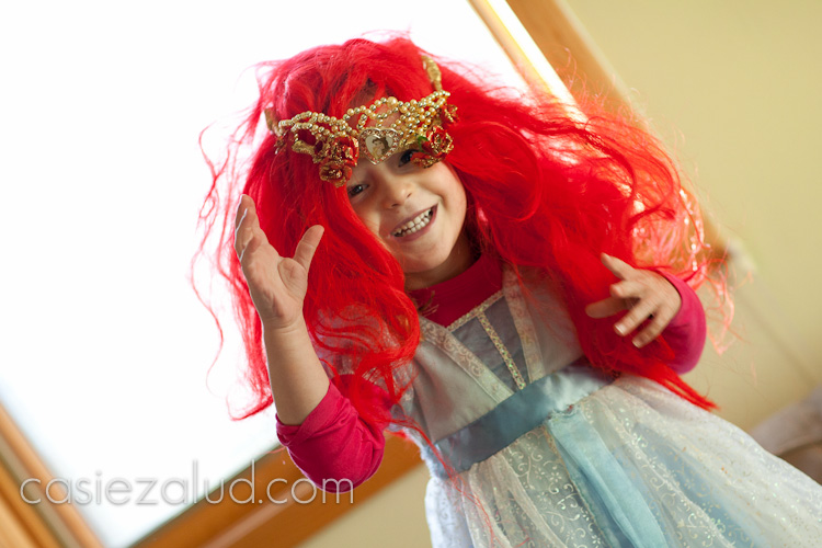 A two year old dressed as Princess Ariel jumping up and down as her red wing and crown falls off and 
