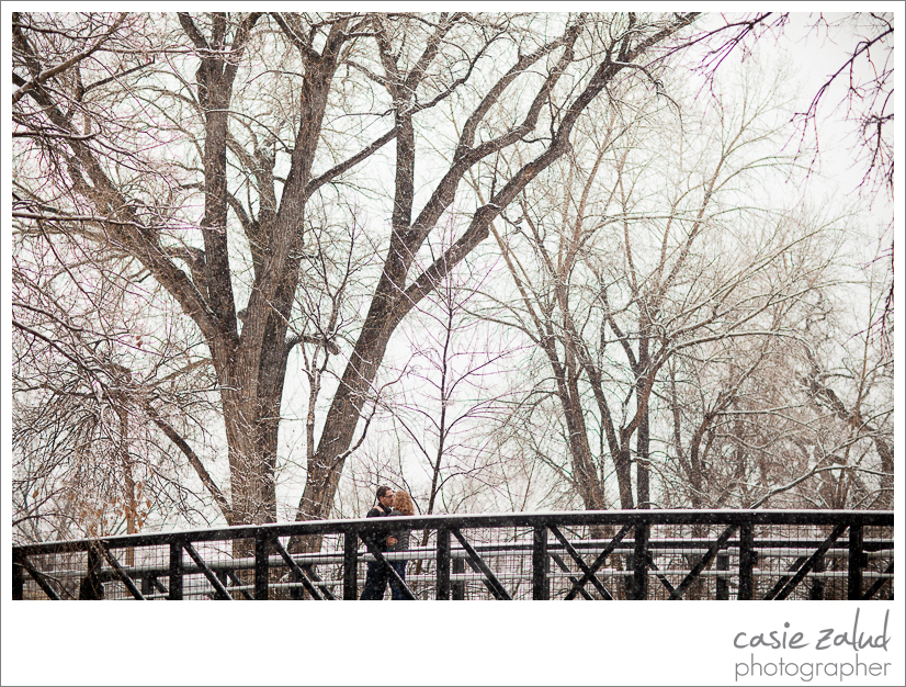 Engaged couple kissing on bridge over Boulder Creek in the snow