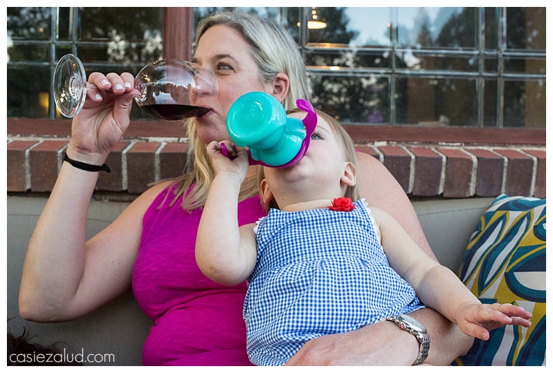 Mom holding a toddler on her lap while she takes a sip of her wine at the same time the toddler sips her milk from her sippie cup