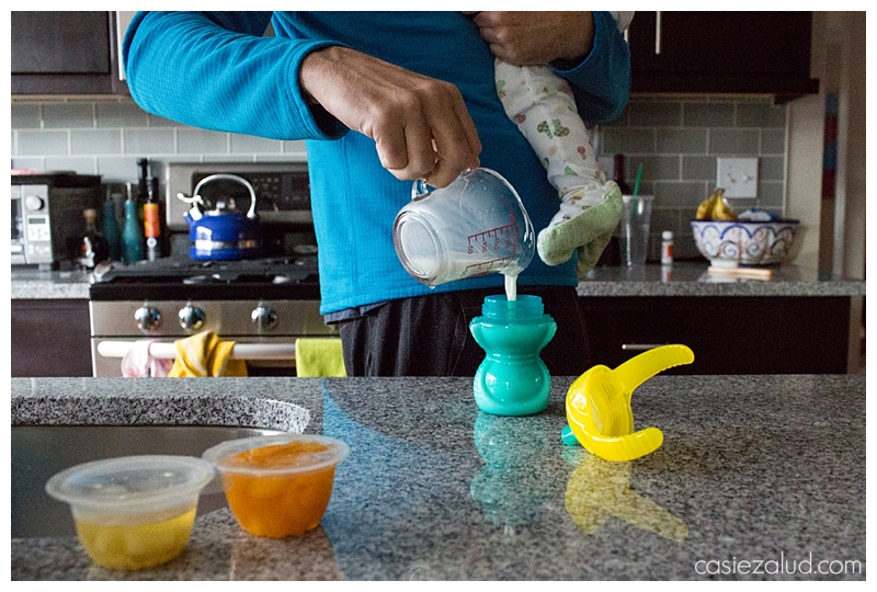 hands of a father holding a babe in a sleeper, while pouring milk into a sippie cup