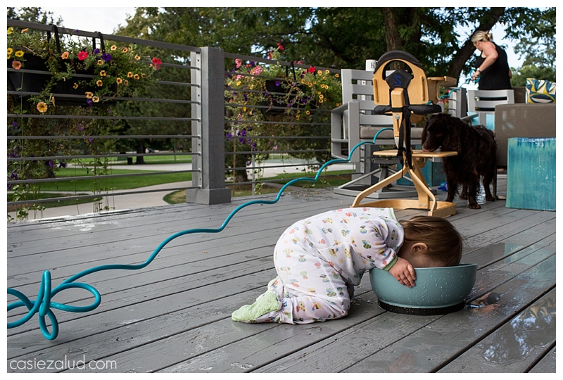 Toddler with her face in a dog bowl, while the dog cleans her high chair and mom waters plants in the background