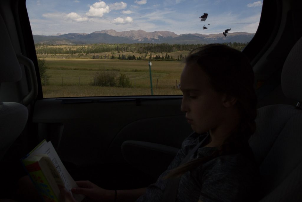 small child in back seat of moving car reading a book as the rocky mountains pass her by